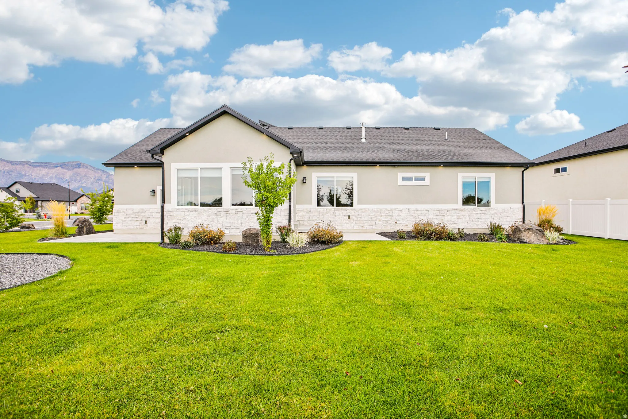 Stucco and stone exterior, dark shingle roof, established landscaping, white vinyl privacy fencing, and mountain backdrop