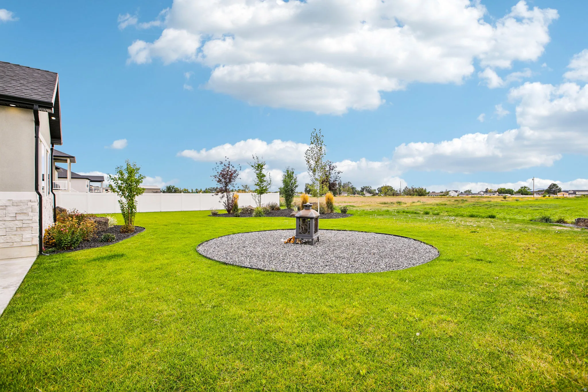Expansive lawn featuring a circular gravel patio with a built-in fire pit, mature landscaping, and a privacy fence