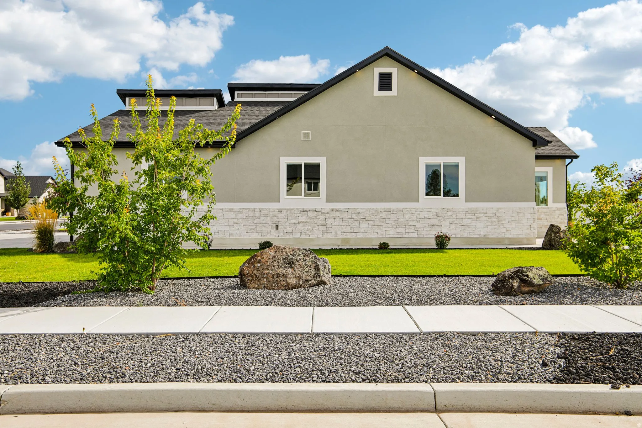Stucco and stone exterior with a black shingle roof, landscaping featuring manicured lawn and decorative rocks, and a concrete sidewalk with gravel beds