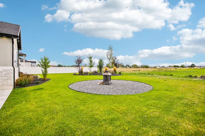 Expansive lawn featuring a circular gravel patio with a built-in fire pit, mature landscaping, and a privacy fence