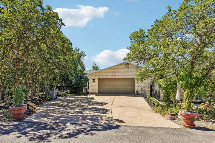 Brick exterior with an attached garage, paved driveway, mature trees, and landscaped front yard