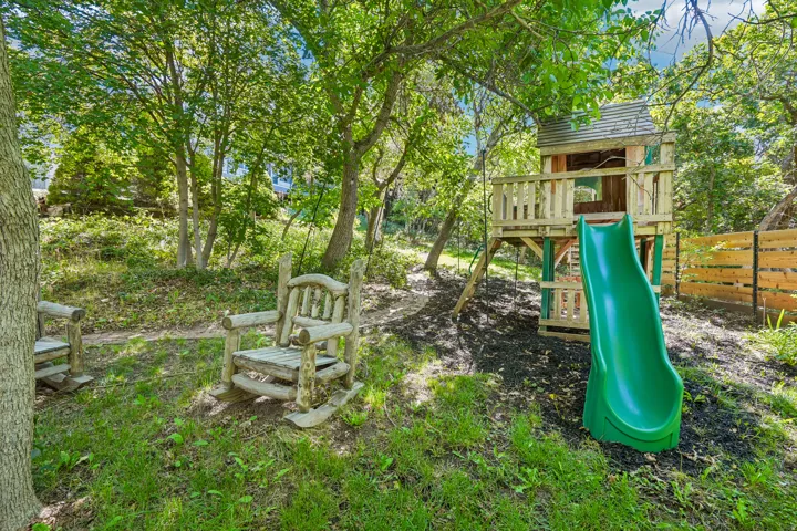 Wood-finished play structure with a green slide, surrounded by mature trees and natural ground cover