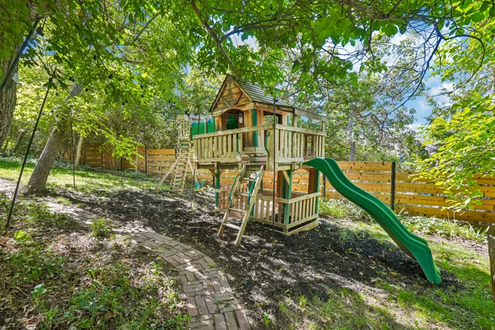 Expansive backyard featuring a custom wooden play structure with a slide, a brick pathway, and mature trees providing natural shade