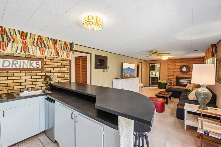 Built-in bar with sink and refrigerator, brick accent wall, fireplace with brick surround, and ceiling fan