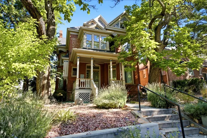 Brick exterior with decorative trim, covered porch with white columns, concrete walkway with handrails, established trees, and varied landscaping