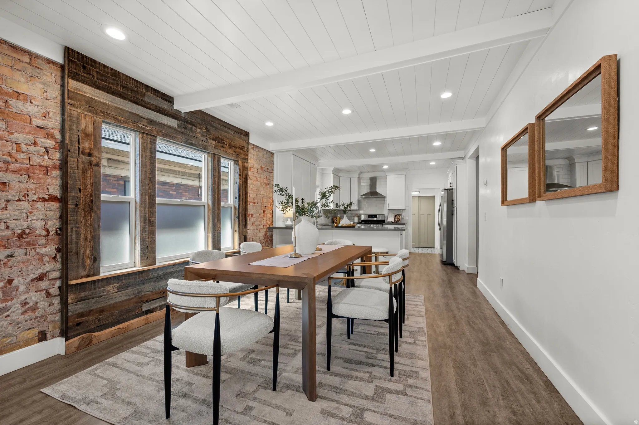 Exposed brick wall, wood-finish flooring, white shiplap ceiling with recessed lighting, and a kitchen featuring a stainless steel range hood