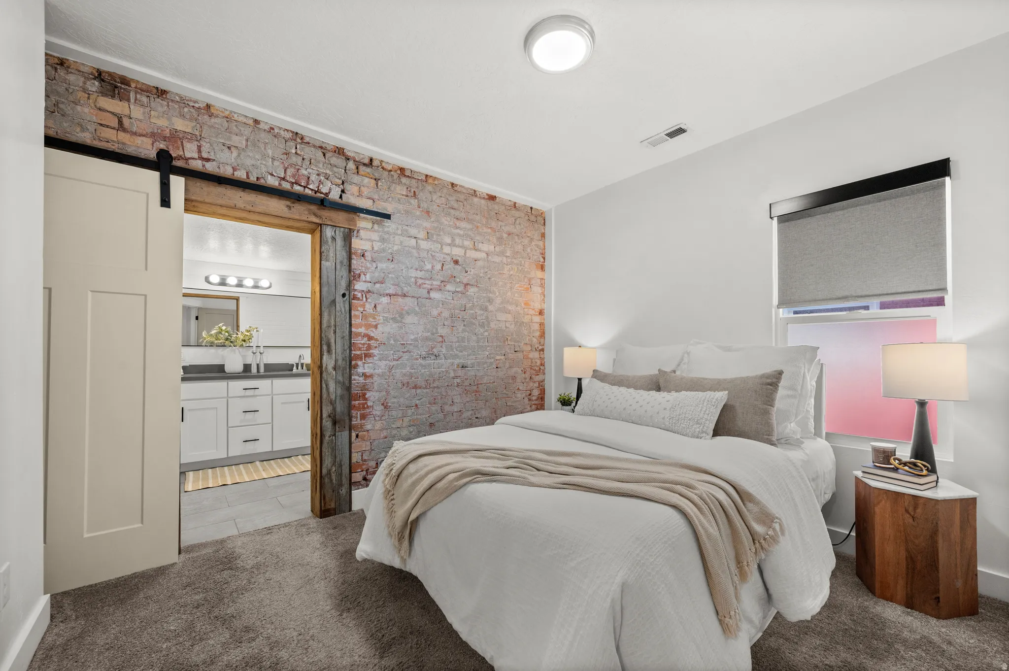 Exposed brick accent wall, sliding barn door, recessed lighting, wall-to-wall carpeting, and a window with a roller shade