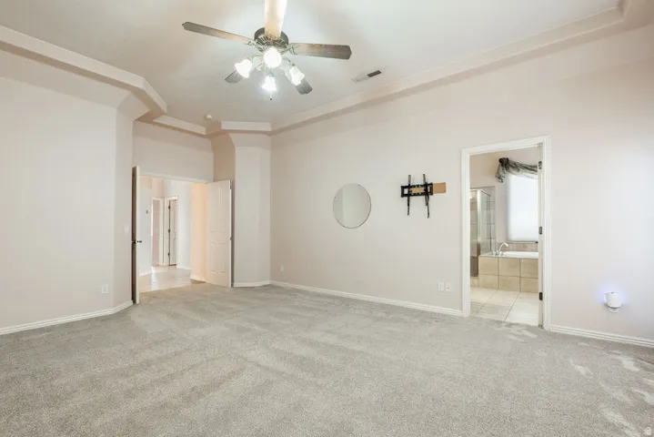 Tray ceiling with crown molding, ceiling fan, carpeted flooring, and a tiled bathroom with a built-in tub