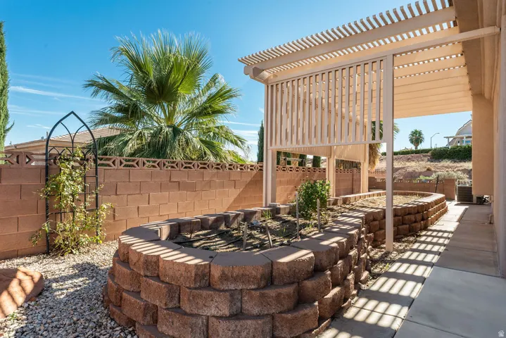 Outdoor living area featuring a slatted pergola, concrete paver patio, block wall fencing, and raised garden beds