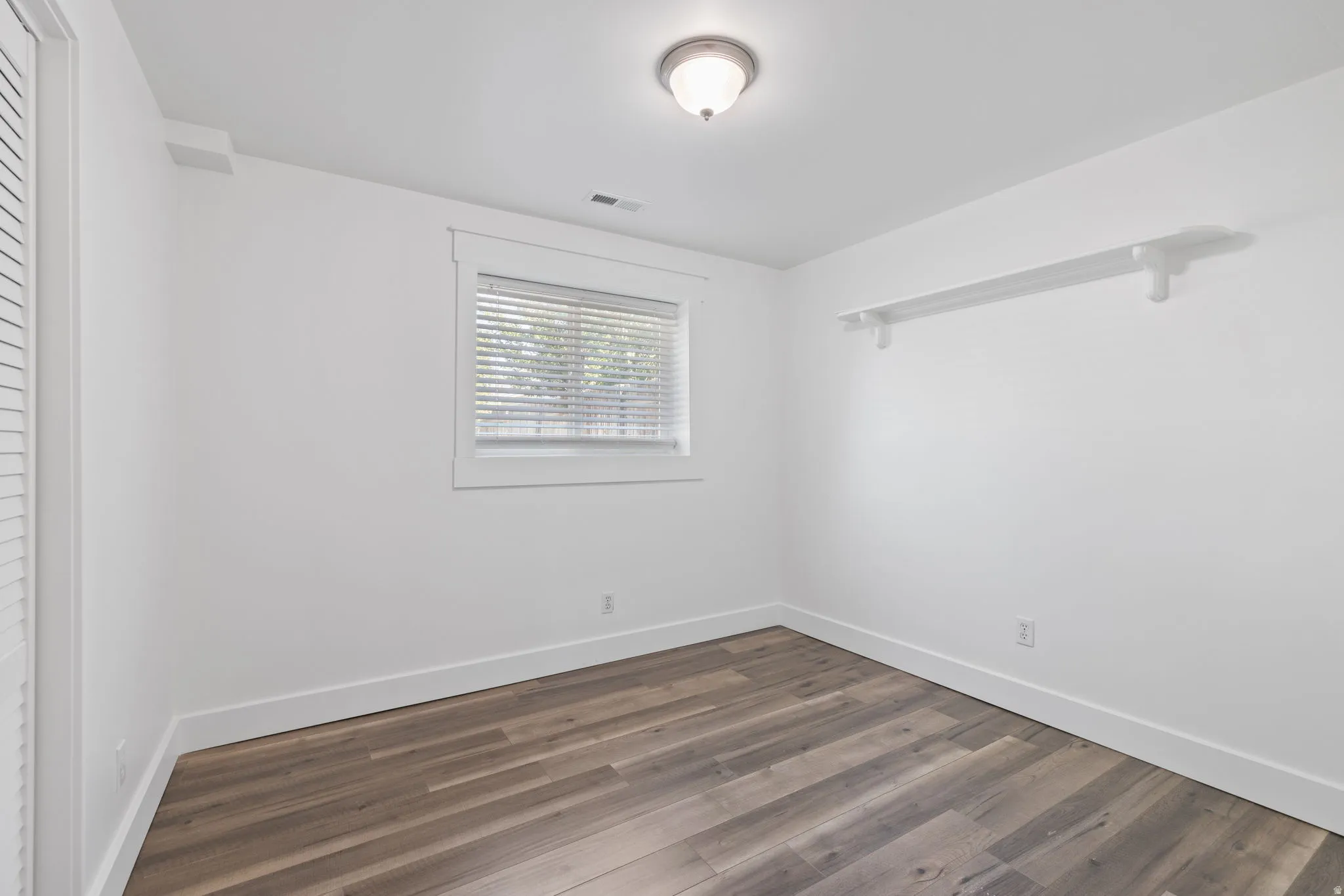 White walls, wood-finish flooring, flush mount ceiling light fixture, window with blinds, and built-in wall shelving