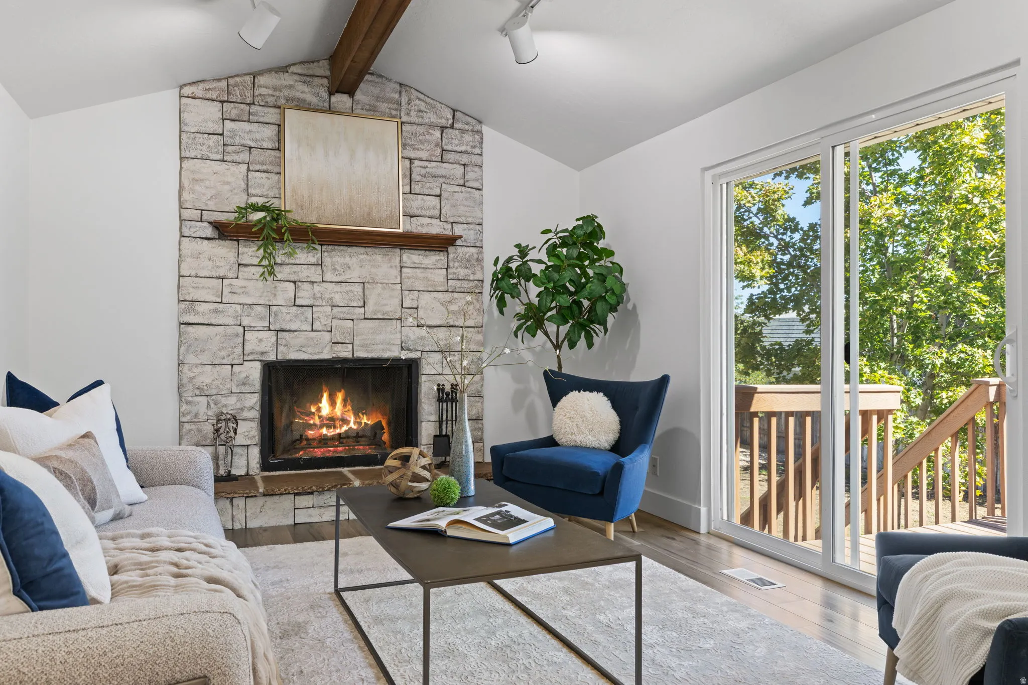 Vaulted ceiling with exposed wood beam, stone-surround fireplace with wood mantel, light wood-finish flooring, and sliding glass door with outdoor access
