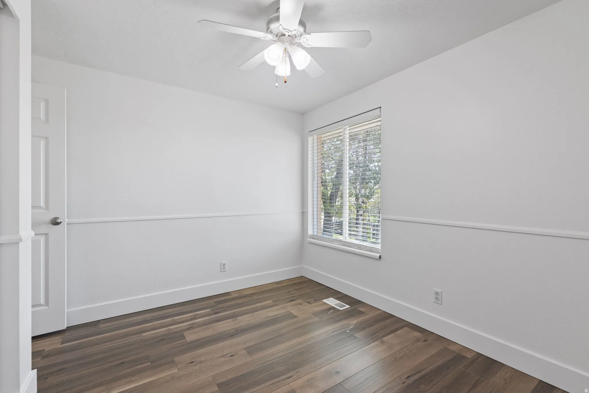 Wood-finish flooring, white walls with chair rail molding, window with blinds, ceiling fan with integrated lighting, and a paneled door