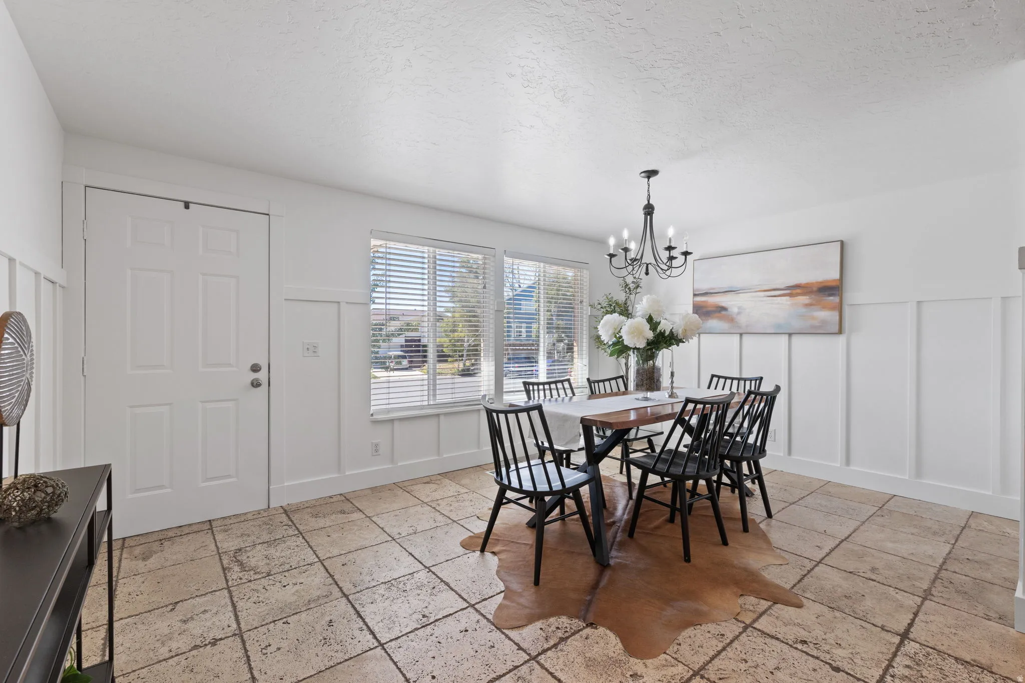 White paneled door, large windows with blinds, board and batten wall detailing, stone tile flooring, and a chandelier with candelabra bulbs