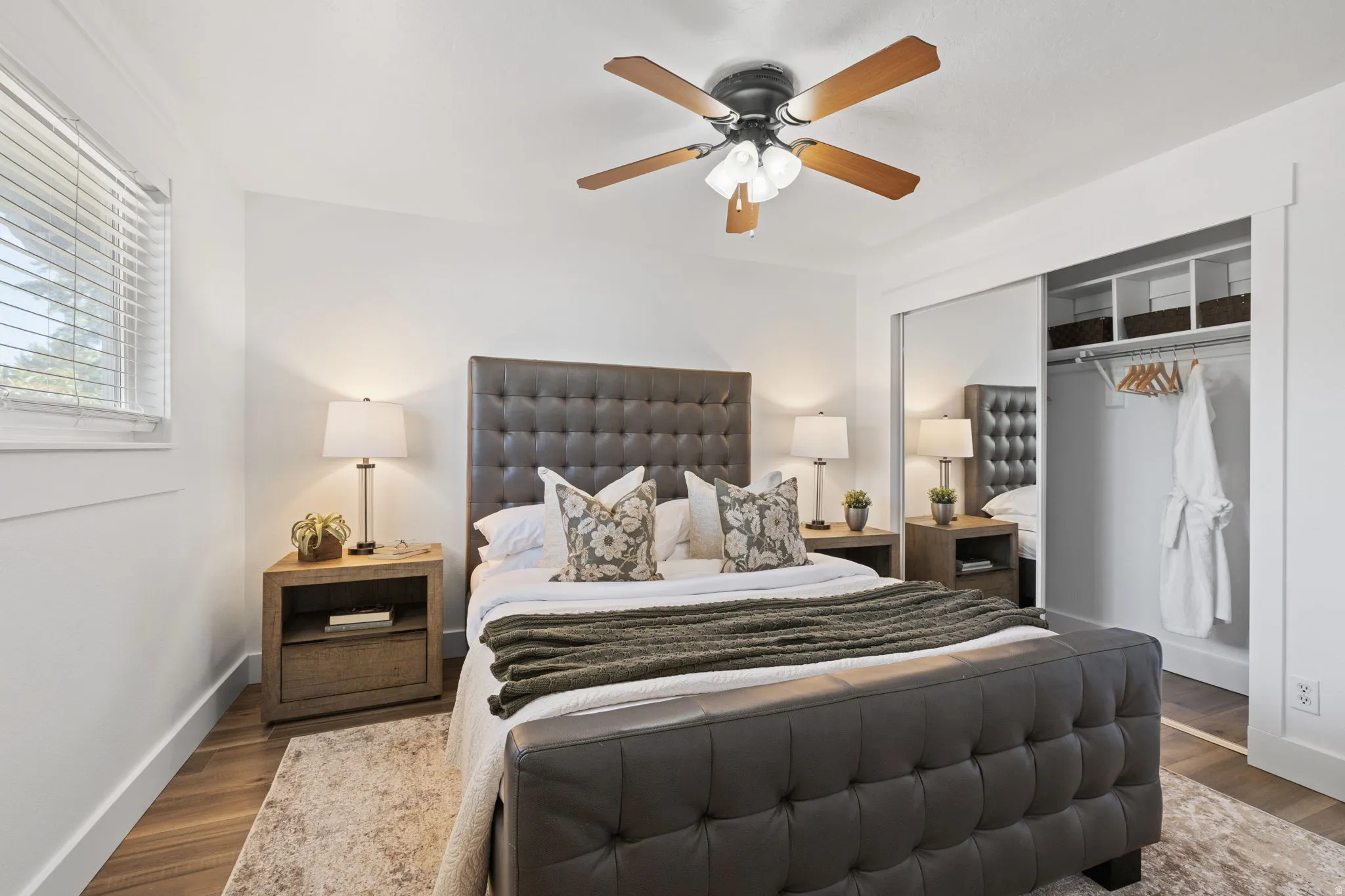 Wood-finish flooring, built-in closet shelving, window with blinds, and a ceiling fan with integrated lighting
