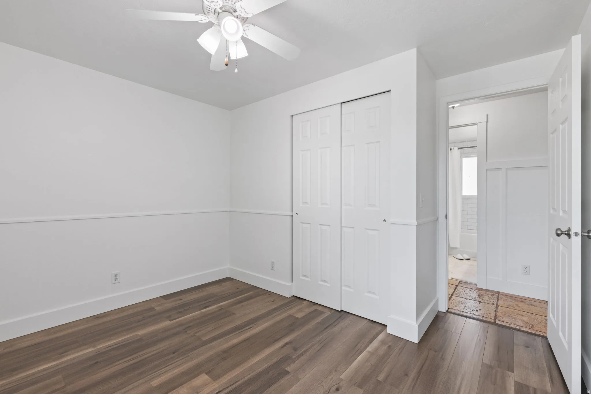 Wood-finish flooring, white walls with wainscoting detail, ceiling fan with light fixture, and white bi-fold closet doors