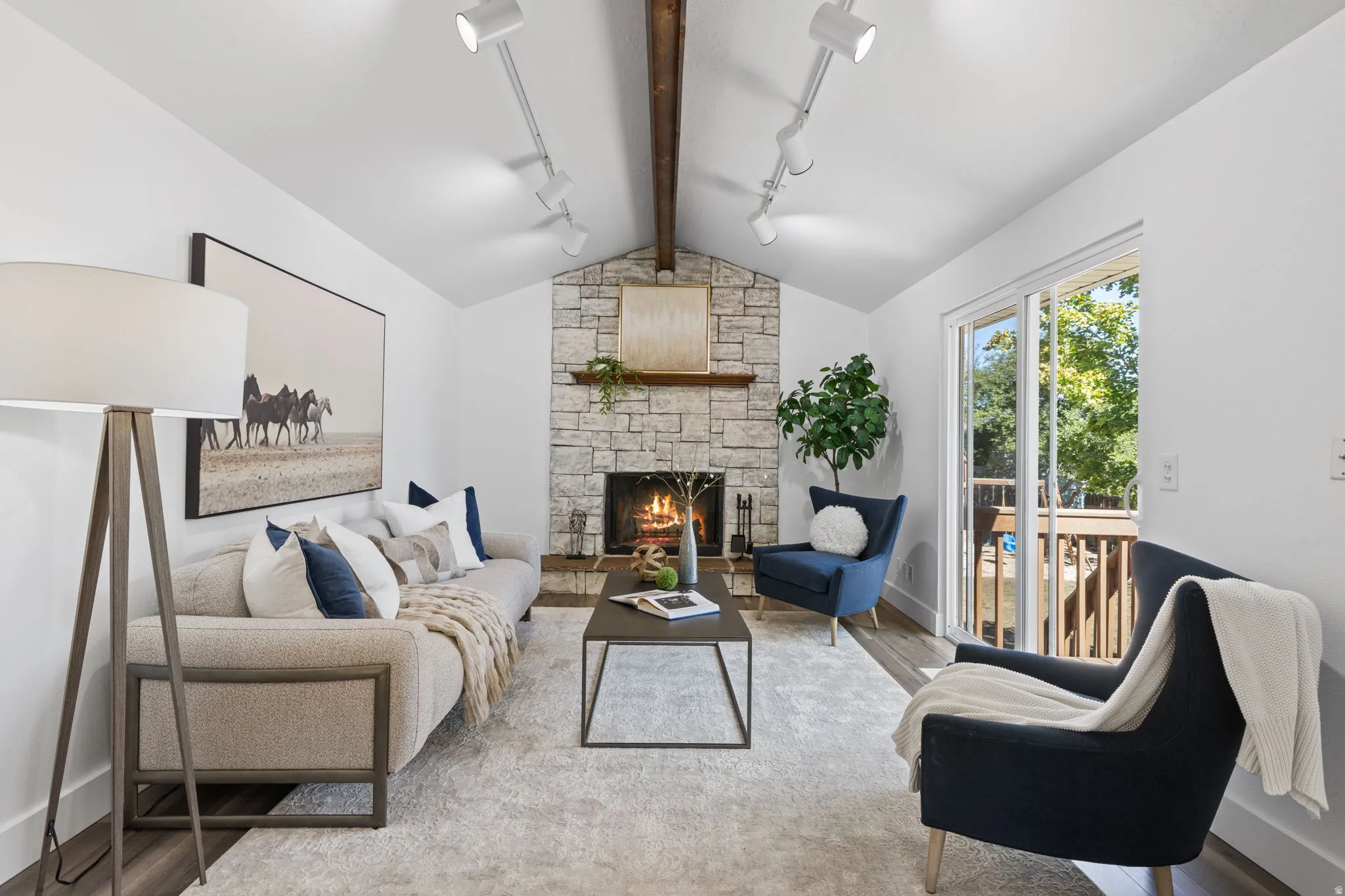 Vaulted ceiling with exposed wood beam, stone-surround fireplace, wood-finish flooring, sliding glass door, and recessed track lighting