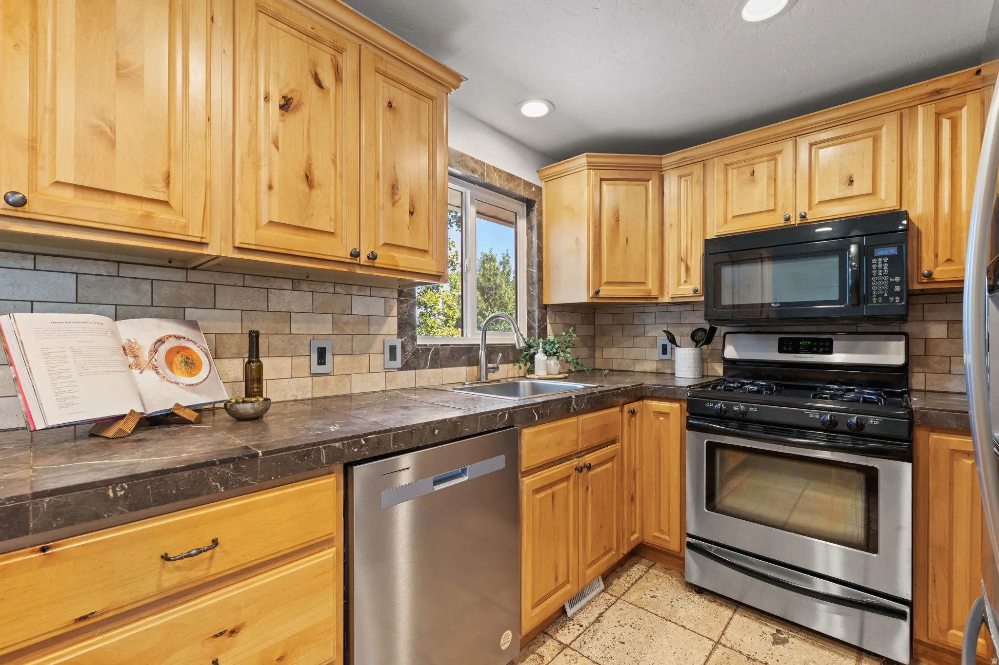 Wood cabinetry with dark hardware, dark countertops, tiled backsplash, stainless steel appliances, and a double basin sink