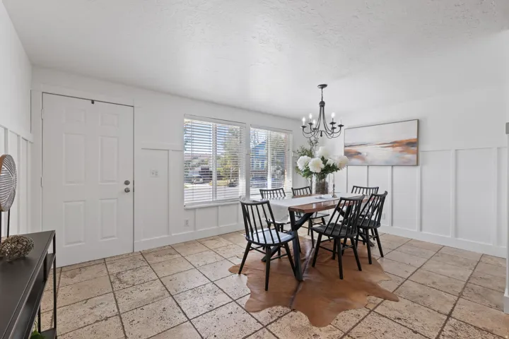 White paneled door, large windows with blinds, board and batten wall detailing, stone tile flooring, and a chandelier with candelabra bulbs
