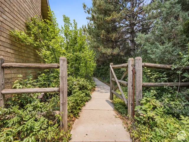Paved walkway with a rustic wood fence and gate, surrounded by mature trees and lush plantings, leading to a brick-clad exterior