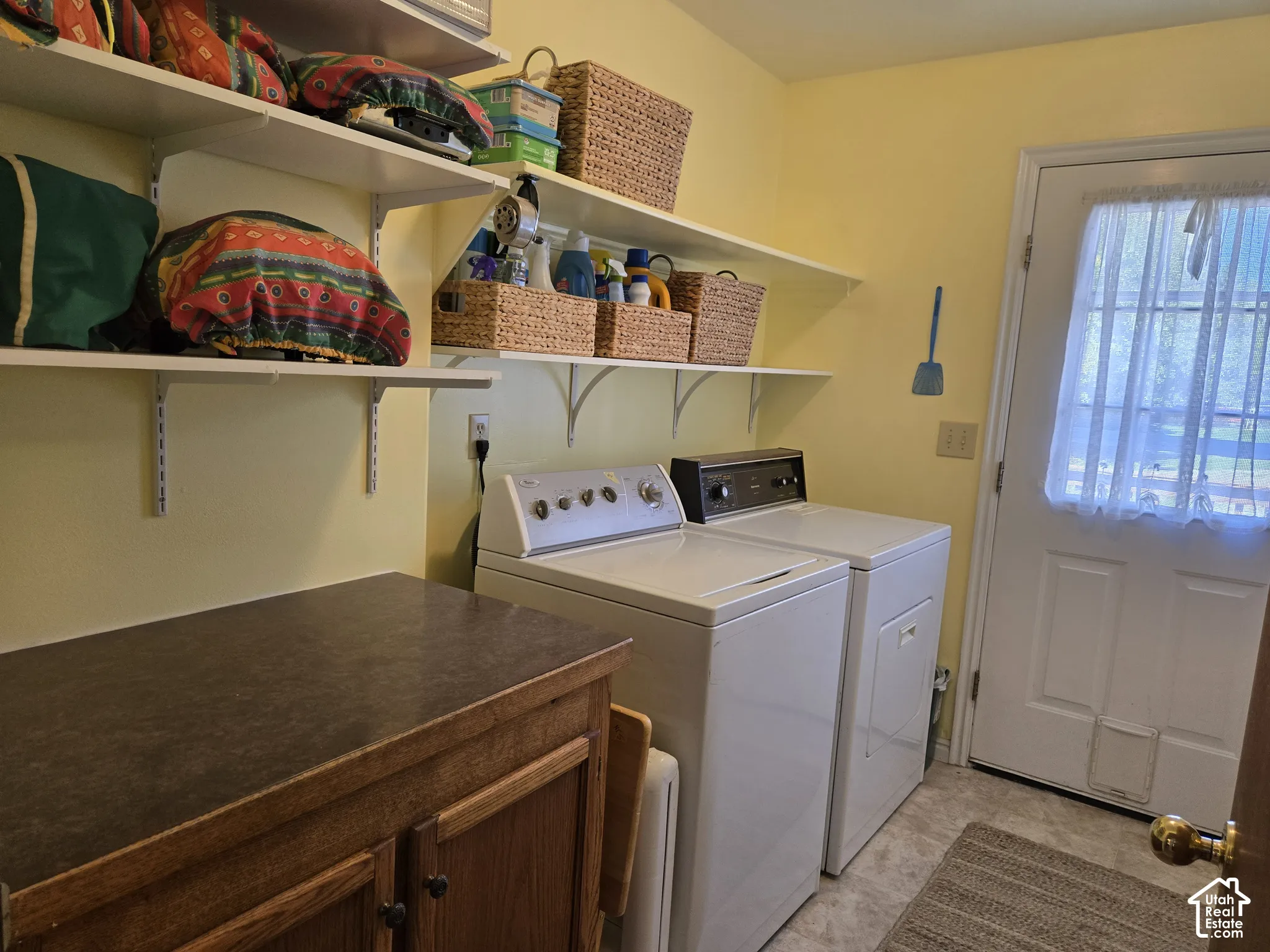 Integrated laundry appliances with durable flooring, open wall shelving, and a built-in wood-finish cabinet with a dark countertop leading to the back yard and ramp