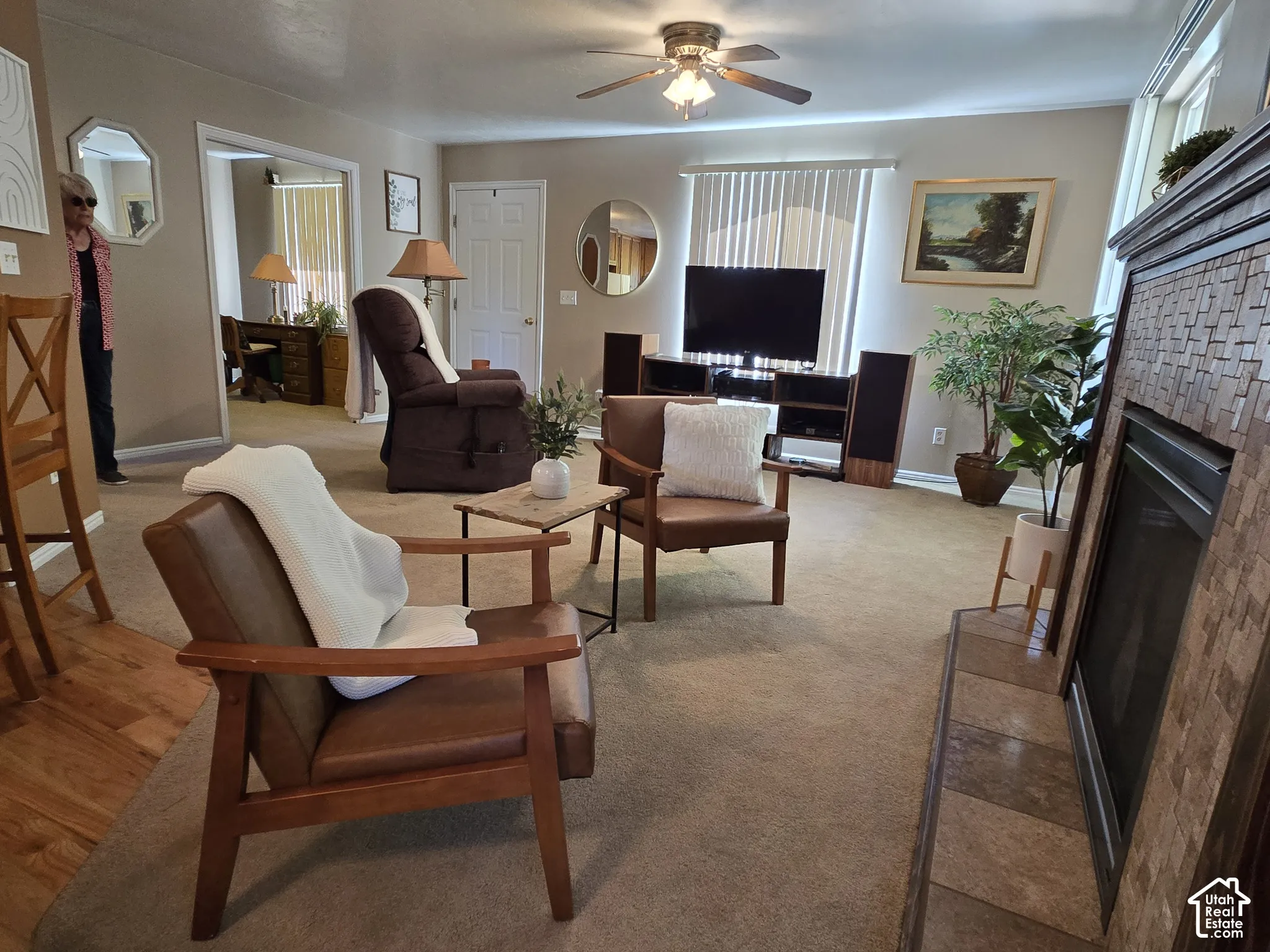 Living area featuring a ceiling fan, fireplace with a tiled surround, carpeted flooring, and an entryway.