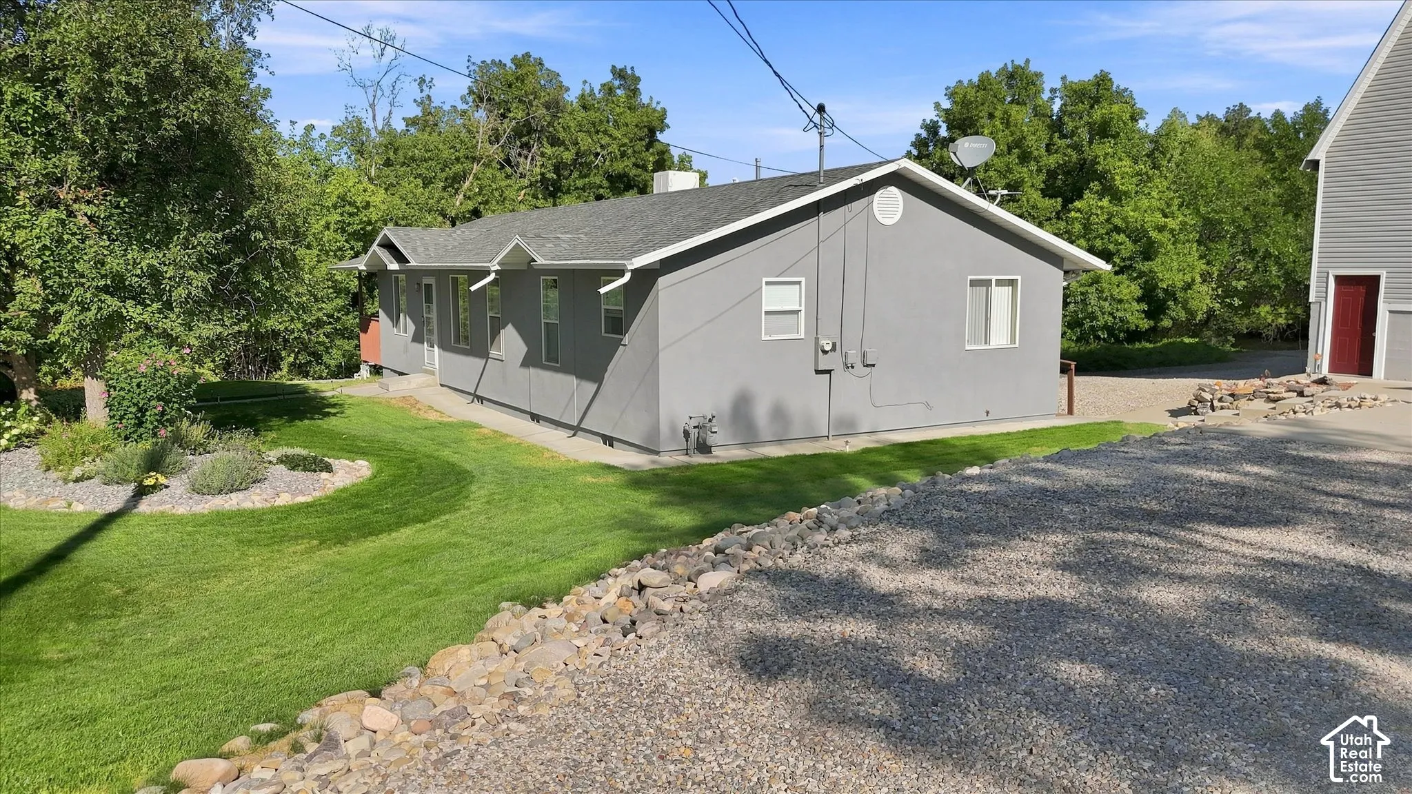 Gray exterior siding, gabled roofline, graveled driveway, manicured lawn with landscaping, and a red entry door on the barn