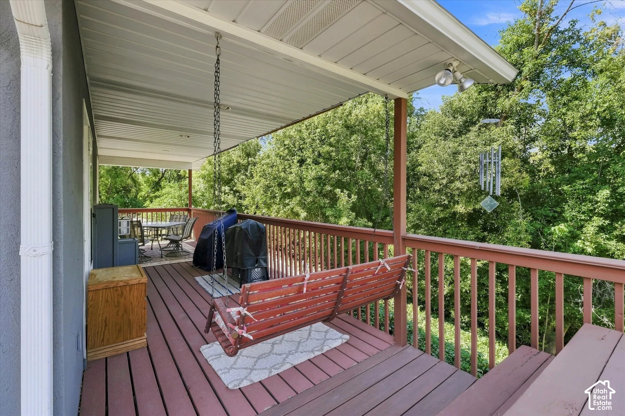 Covered porch featuring a red wood-finish deck and railing, with lush green trees providing a natural backdrop