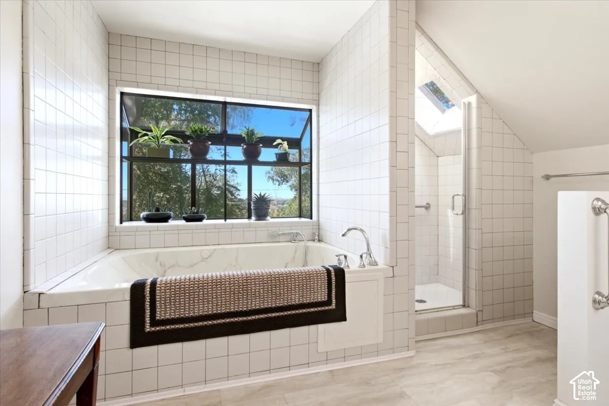 Built-in soaking tub with deck-mounted fixtures, separate shower with glass enclosure, white square tile surround, skylight, and large window