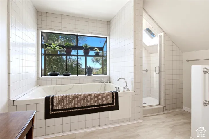 Built-in soaking tub with deck-mounted fixtures, separate shower with glass enclosure, white square tile surround, skylight, and large window