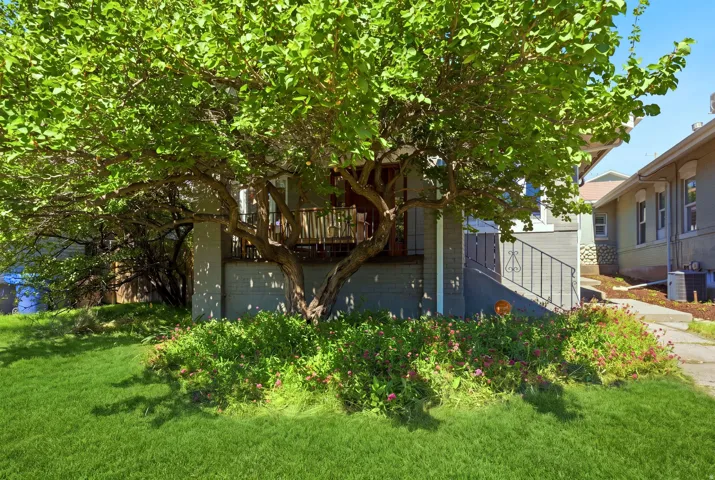 Manicured lawn with mature tree coverage, front porch with wood-finish railing, painted exterior siding, and wrought iron handrail