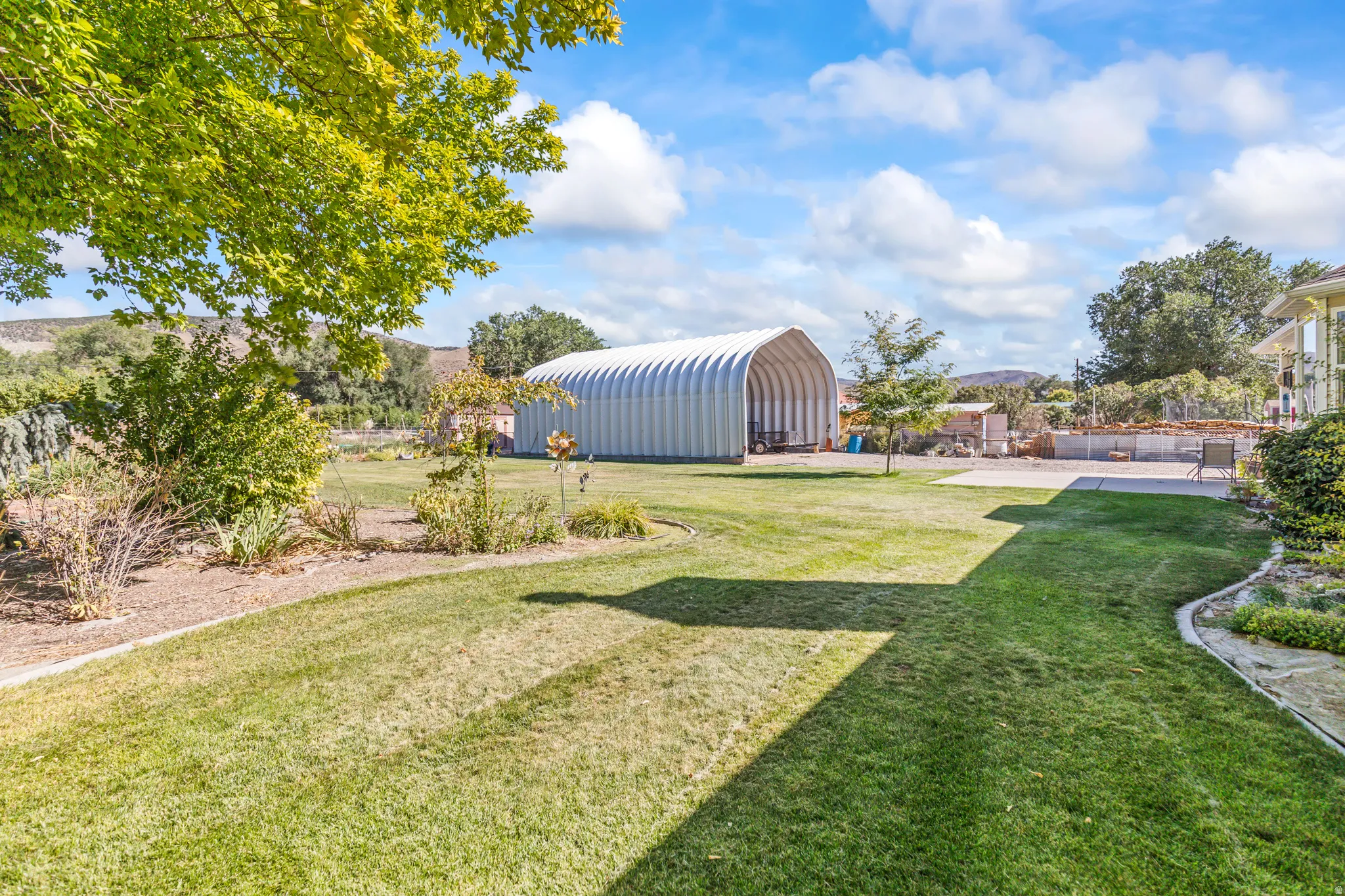 Expansive lawn area with a large arch-style outbuilding, mature trees, landscaped garden beds, and a paved patio area