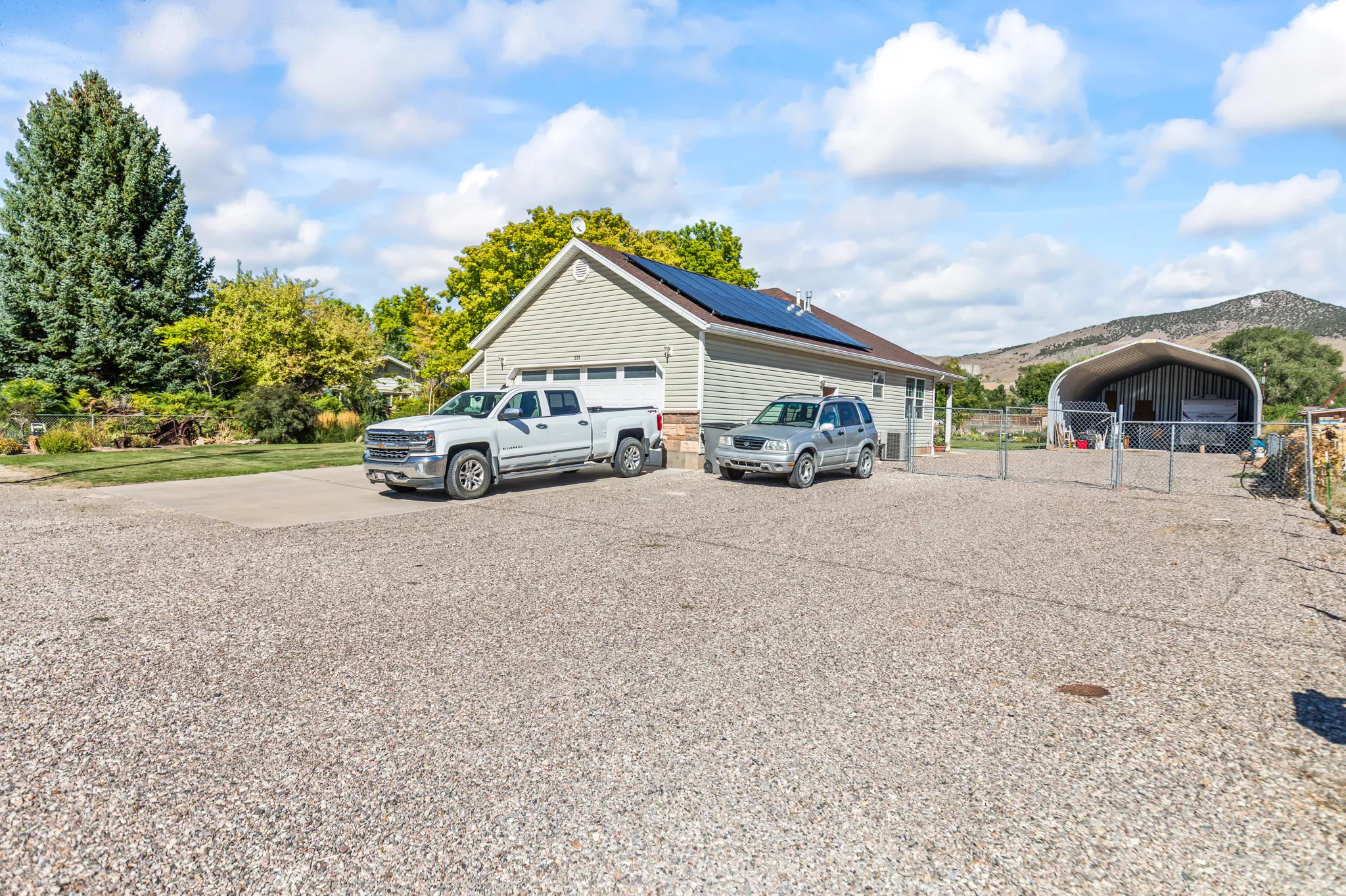 Exterior featuring light-colored siding, a two-car garage, roof-mounted solar panels, a gravel driveway, and a metal-frame utility structure