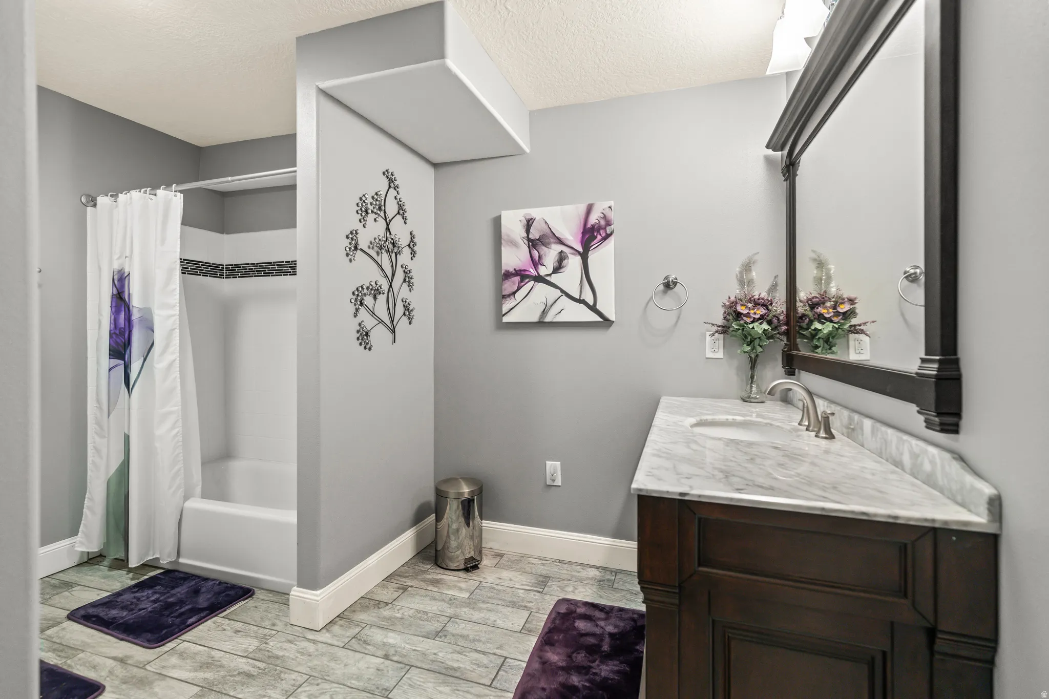 Wood-finish plank flooring, single vanity with integrated sink, stone countertop, built-in tub with tile surround, and an ornate framed mirror