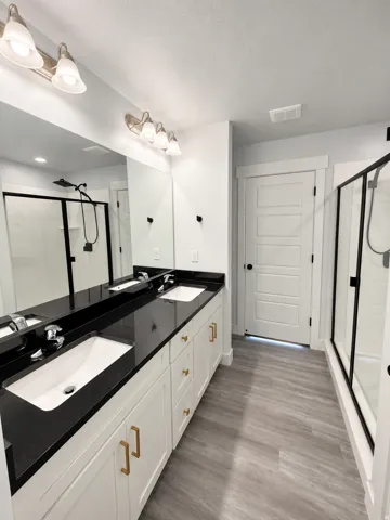 Dual vanity with integrated sinks and dark countertops, brushed nickel lighting fixture, framed mirror, wood-finish flooring, and a glass-enclosed shower