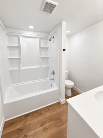White bathtub with integrated shelving, chrome shower fixtures, wood-finish flooring, white vanity with integrated sink, and recessed lighting