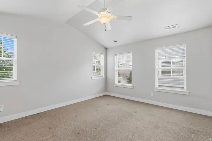 Vaulted ceiling, ceiling fan with light fixture, multiple windows with blinds, and neutral carpeting