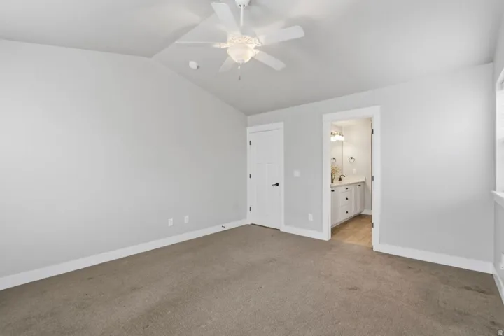 Vaulted ceiling with a ceiling fan, neutral wall paint, carpet flooring, a solid panel interior door, and a bathroom featuring a vanity with a countertop and undermount sink