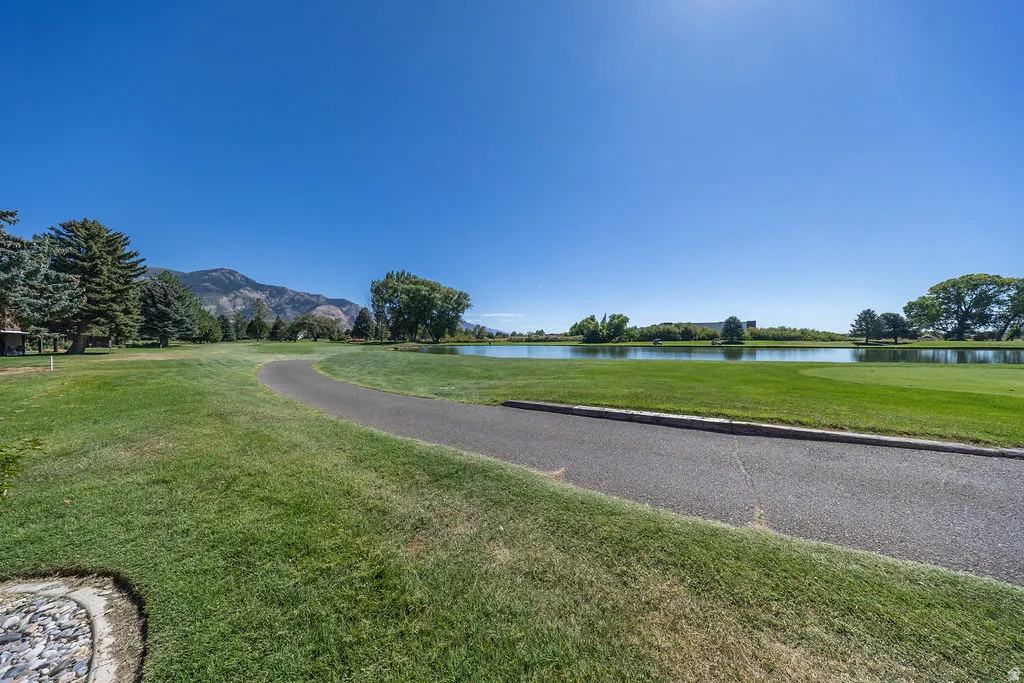 Manicured lawn with a paved pathway, expansive water feature, mature trees, and mountain backdrop