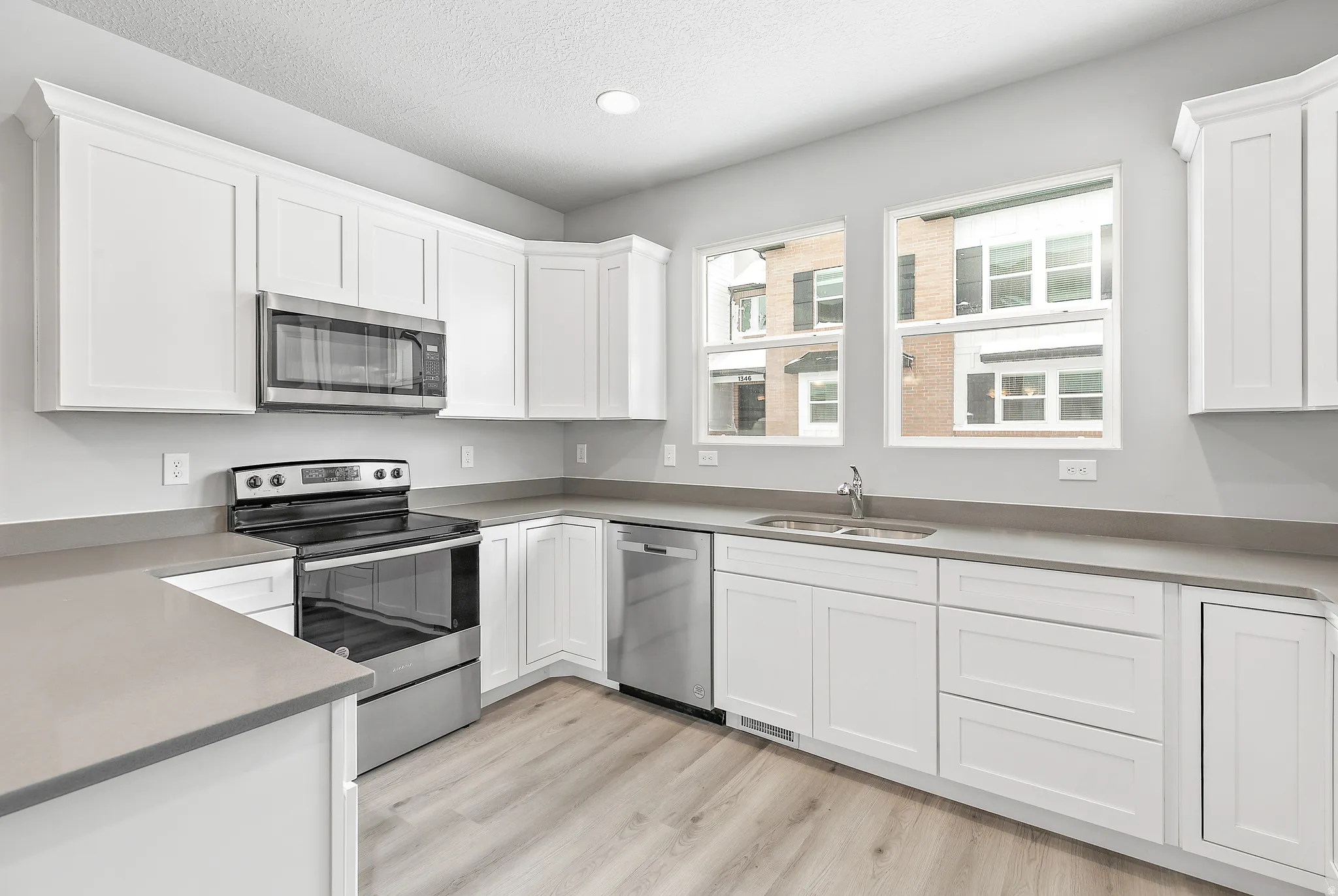 Kitchen featuring white shaker-style cabinetry, stainless steel appliances, light gray countertops, wood-finish flooring, and two double-hung windows