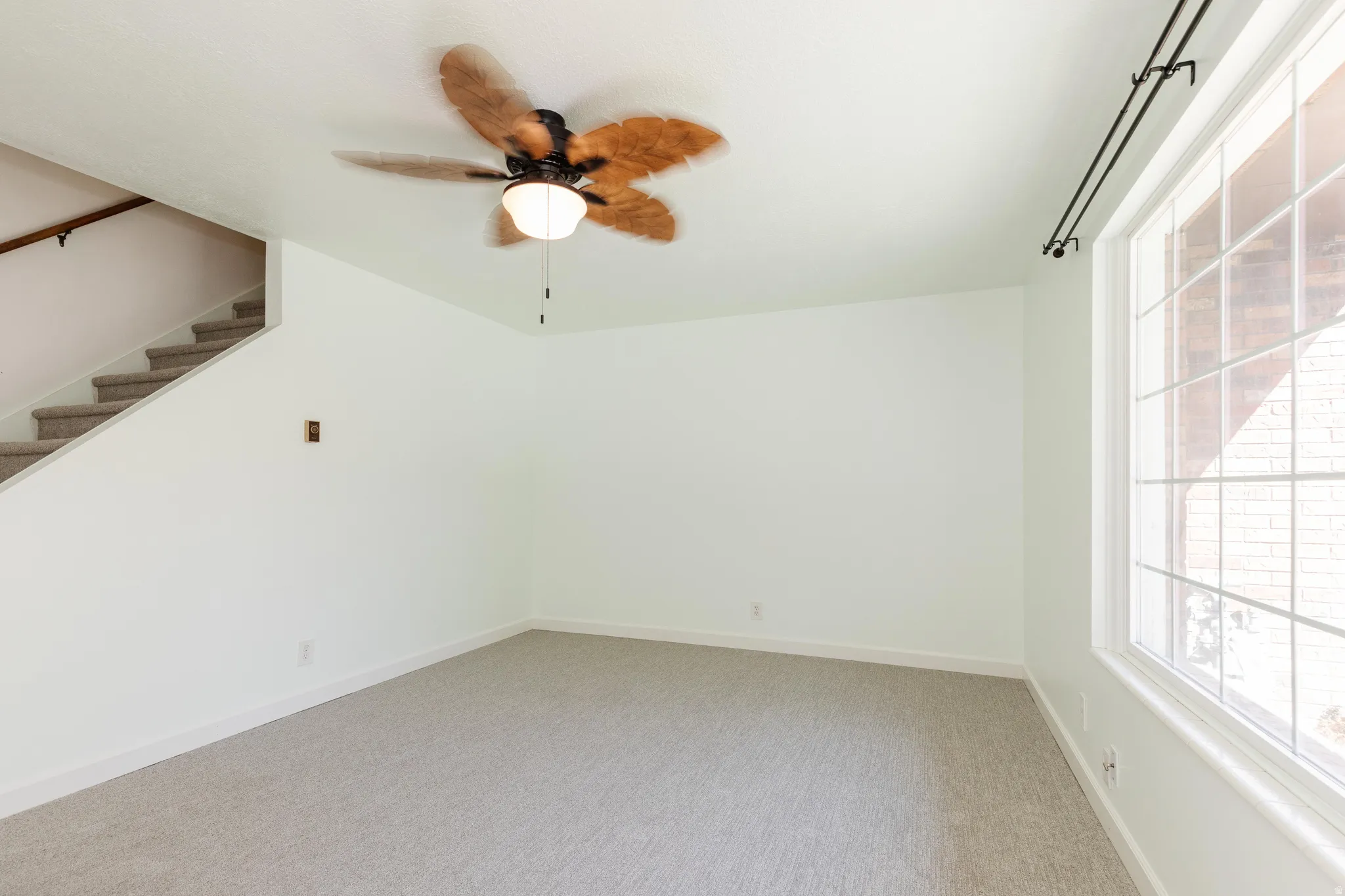 Light-toned carpeting, white walls, ceiling fan with integrated lighting, window with grid pattern, and a staircase with carpeted treads