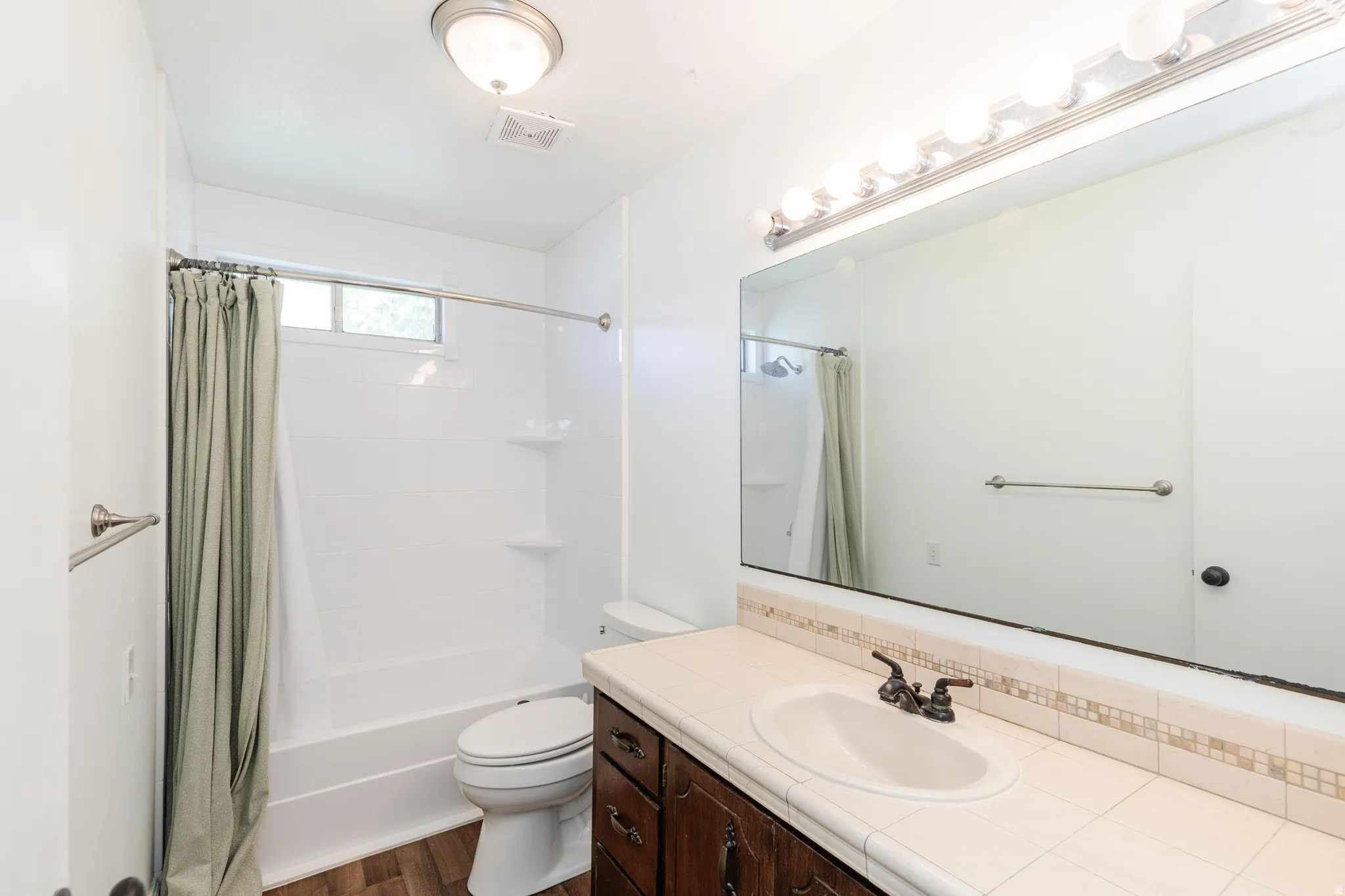 White tub and shower combination with a window, wood-finish flooring, and a vanity featuring an integrated sink, tile countertop, and wood-finish cabinetry
