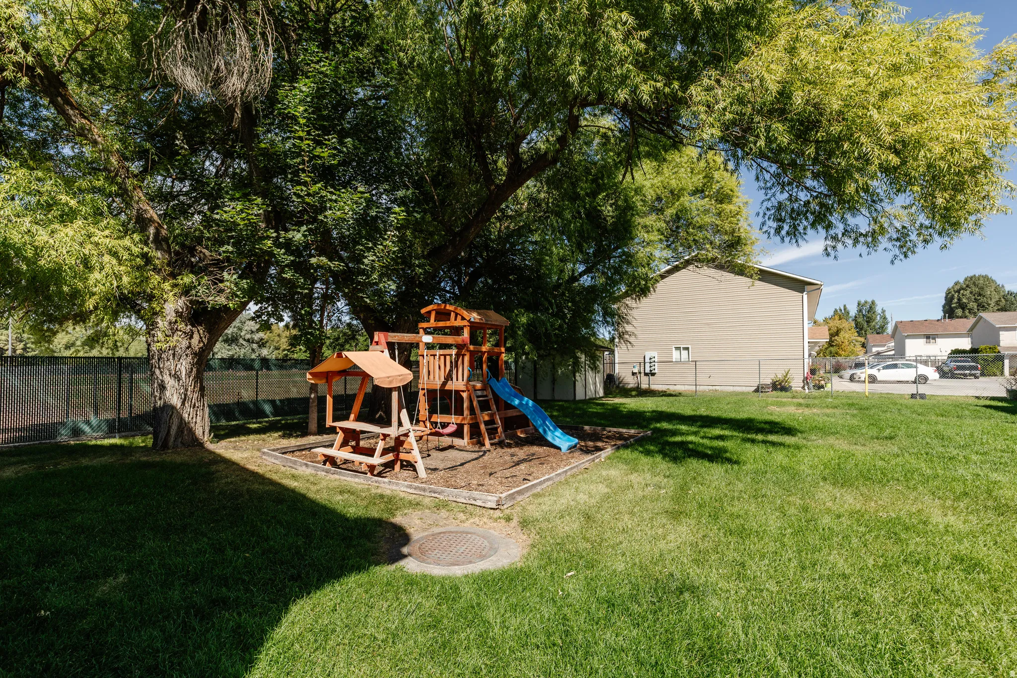 Expansive lawn area featuring a mature tree, a built-in wooden playset with a blue slide, and a chain-link fence