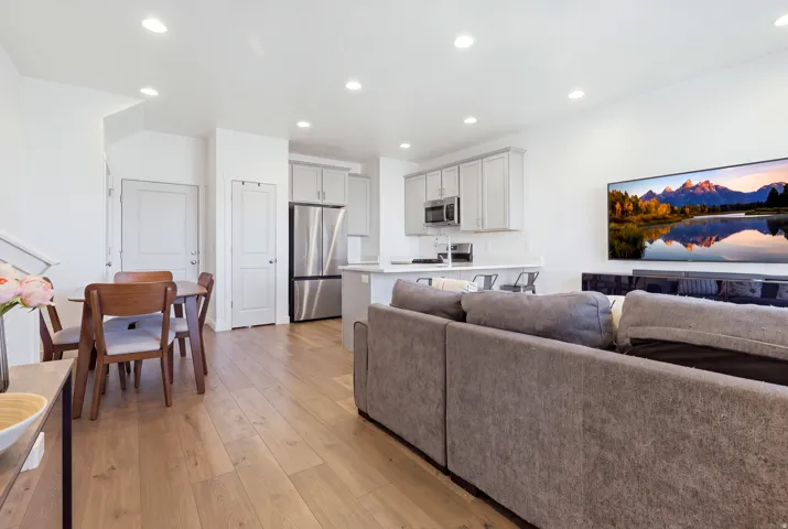 Open-concept living area with wood-finish flooring, recessed lighting, and a white kitchen featuring light gray cabinetry, stainless steel appliances, and a kitchen island with seating