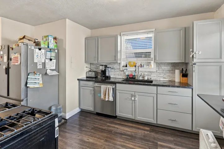 Light gray cabinetry with brushed nickel hardware, dark countertops, tile backsplash, stainless steel dishwasher, and wood-finish flooring