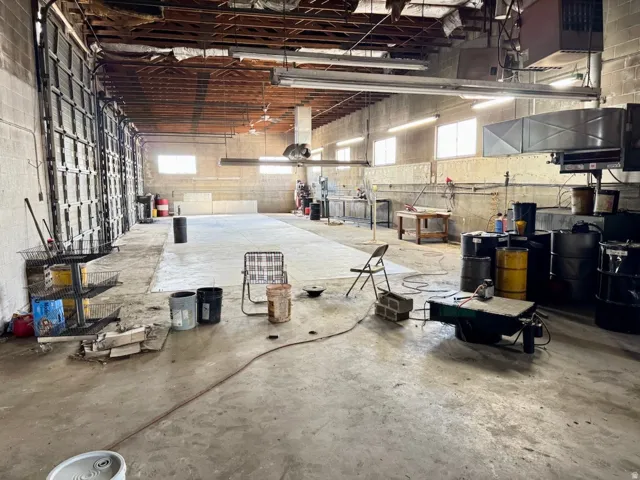 Spacious interior featuring concrete flooring, exposed wood beam ceiling, multiple roll-up doors, high windows, and overhead fluorescent lighting