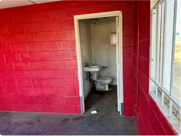 Red-painted cinder block exterior, white-framed window, and a white-framed doorway opening to a utility sink and toilet