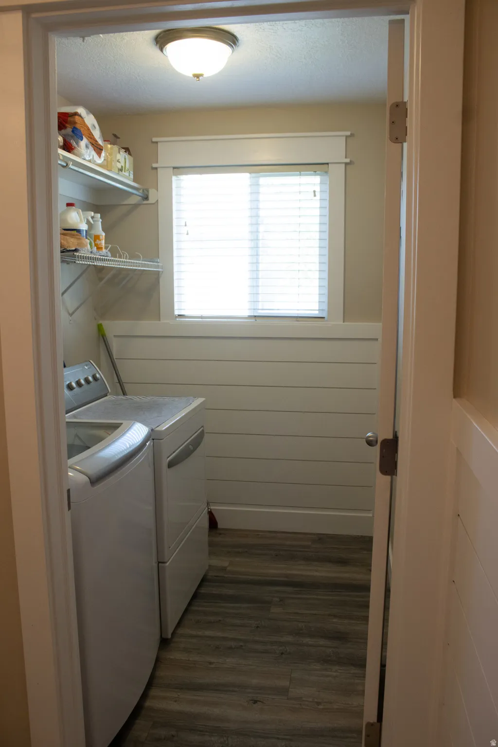 Wood-finish flooring, white shiplap wall accent, window with white blinds, flush mount ceiling light, and built-in wire shelving