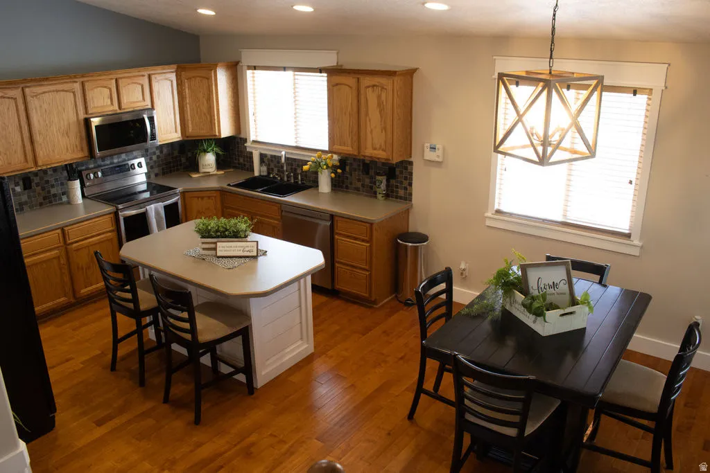 Hardwood flooring throughout, L-shaped kitchen cabinetry, kitchen island with seating, stainless steel appliances, and a decorative pendant light fixture