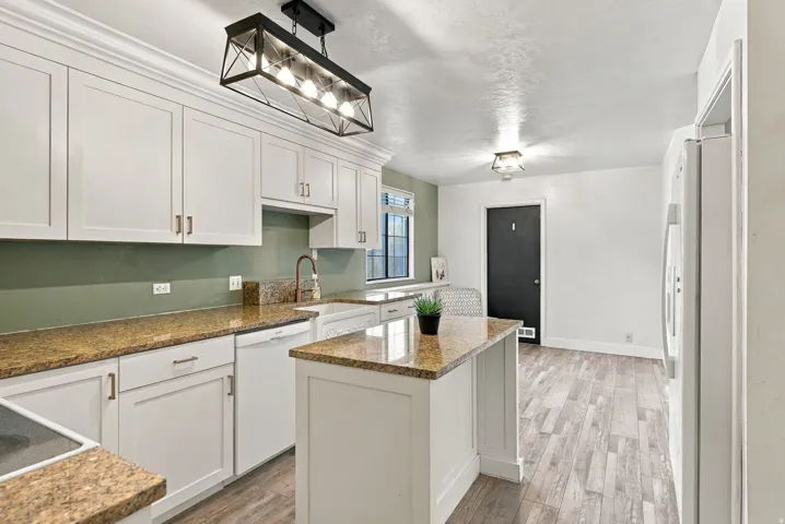 White shaker-style cabinetry with crown molding, granite-finish countertops, wood-finish flooring, and an island with a built-in sink