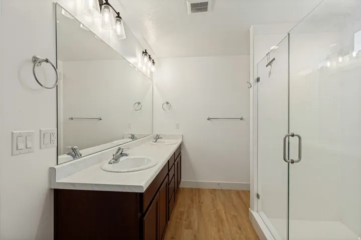 Dual vanity with integrated sinks, wood-finish flooring, full-width mirror, brushed nickel fixtures, and a glass-enclosed shower